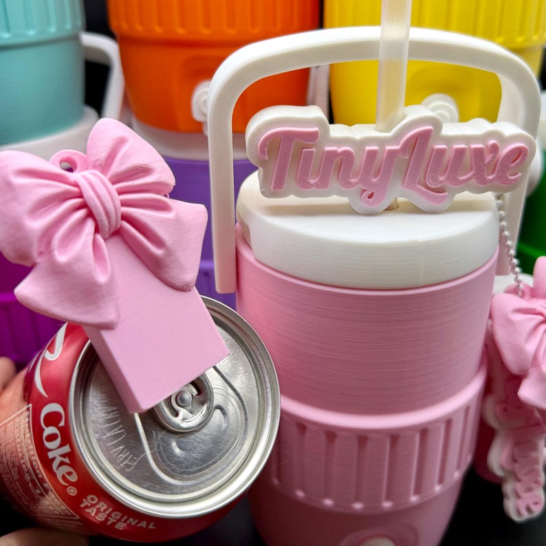 May include: A pink and white Tiny Luxe beverage container with a white handle and a straw. A pink bow-shaped can tab puller is on a Coke can. Other colorful containers are in the background.