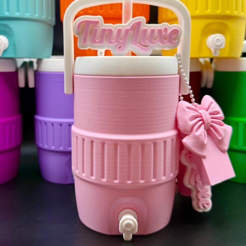 May include: A pink water cooler with a white handle and a white top. The cooler has a spigot and a pink bow-shaped charm with the text "Tiny Luxe". Other coolers in various colors are in the background.