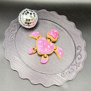 May include: A whimsical, heart-shaped cookie-themed fidget spinner with pink frosting and sprinkles, surrounded by a decorative plate and a disco ball. The spinner has a tan base with pink frosting and sprinkles. The disco ball is silver and reflective.