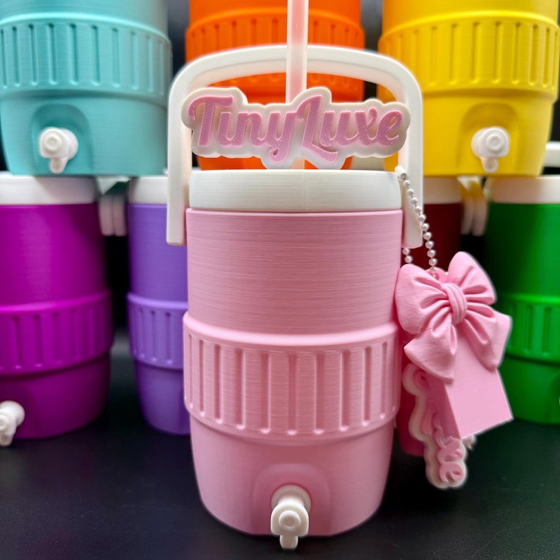 May include: A pink water cooler with a white handle and lid, featuring the text "Tiny Luxe". It has a white spigot and a pink bow-shaped tag. Other coolers in various colors are in the background.