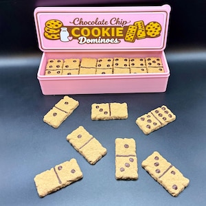 Puede incluir: Una caja rosa de domin&oacute; de galletas con chispas de chocolate. La tapa de la caja presenta las palabras "Chocolate Chip COOKIE Dominoes". Los domin&oacute;s son de color beige con detalles de chispas de chocolate marr&oacute;n.