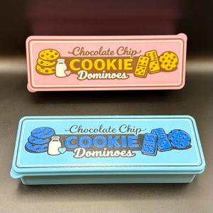 Puede incluir: Se muestran dos cajas rectangulares de "Chocolate Chip Cookie Dominoes". Una es rosa con ilustraciones de galletas y domin&oacute;s amarillos, la otra es azul con ilustraciones de galletas y domin&oacute;s azules. El texto de ambas cajas es cursivo.