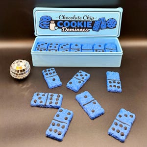 Puede incluir: Una caja azul claro con las palabras "Chocolate Chip Cookie Dominoes" en la tapa, llena de domin&oacute;s azules con detalles de chispas de chocolate. Varios domin&oacute;s est&aacute;n esparcidos alrededor de la caja. Una peque&ntilde;a bola de discoteca est&aacute; a la izquierda.