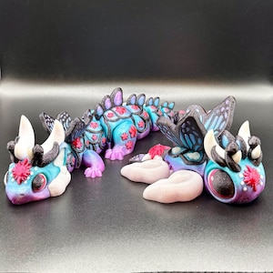 May include: A colorful, articulated dragon figurine with butterfly wings and flower accents. The dragon features a gradient of blue, purple, and pink hues. The figurine has large eyes and is decorated with red flowers. The dragon is a unique, whimsical art piece.