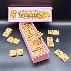Puede incluir: Una caja rectangular rosa llena de domin&oacute;s de galletas con chispas de chocolate. Los domin&oacute;s son de color marr&oacute;n claro con chispas de chocolate marr&oacute;n oscuro y puntos negros. Una tapa rosa con las palabras "Chocolate Chip COOKIE Dominoes" est&aacute; encima de la caja.