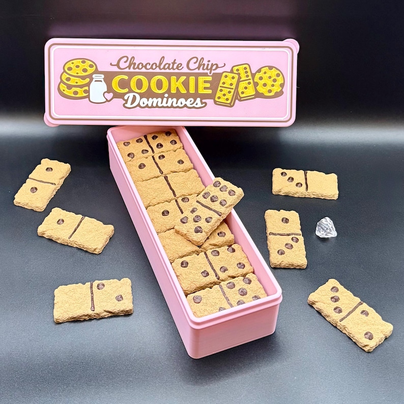 Puede incluir: Una caja rectangular rosa llena de domin&oacute;s de galletas con chispas de chocolate. Los domin&oacute;s son de color marr&oacute;n claro con chispas de chocolate marr&oacute;n oscuro y puntos negros. Una tapa rosa con las palabras "Chocolate Chip COOKIE Dominoes" est&aacute; encima de la caja.