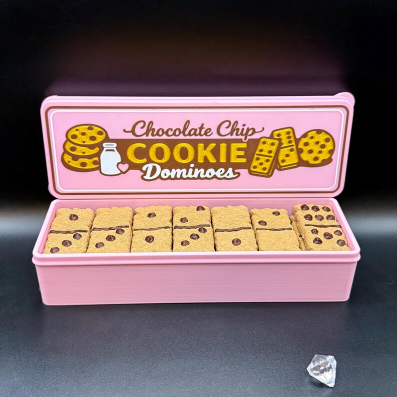 Puede incluir: Una caja rectangular rosa con tapa que muestra las palabras "Chocolate Chip Cookie Dominoes". Dentro, domin&oacute;s que parecen galletas con chispas de chocolate. La tapa tiene ilustraciones de galletas y una botella de leche.