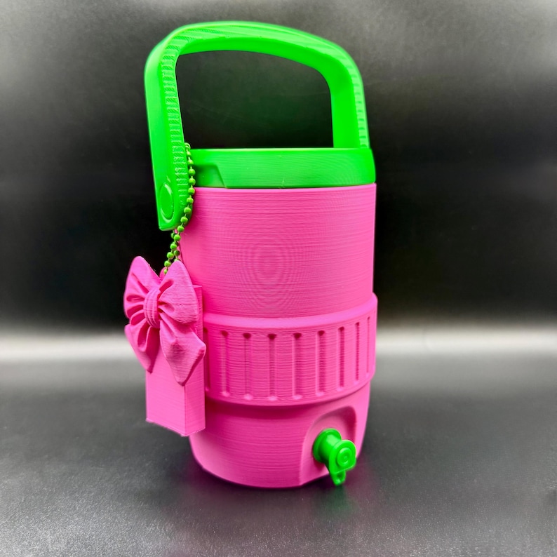 May include: A bright pink water cooler with a green handle, lid, and spigot. A pink bow and tag are attached to the side with a green beaded chain. The cooler has a ribbed design around the middle and bottom.