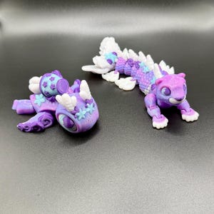 May include: Two articulated fantasy creatures in shades of purple, blue, and white. One resembles a feline with a long, segmented body and a fin-like tail. The other is a rounder, more abstract design with floral accents.