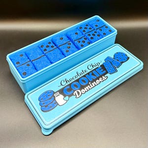 Puede incluir: Una caja azul claro de "Chocolate Chip Cookie Dominoes" con tapa a juego. Los domin&oacute;s son azules con puntos negros. La tapa presenta ilustraciones de galletas y leche.