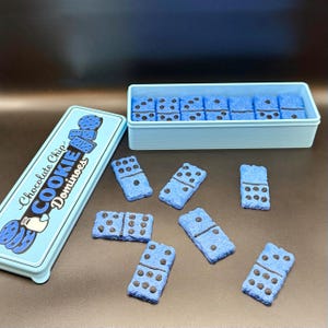 Puede incluir: Una lata y un recipiente azul claro contienen domin&oacute;s con forma de galleta. Los domin&oacute;s son azules con detalles de chispas de chocolate marr&oacute;n oscuro. La lata dice "Chocolate Chip Cookie Dominoes" en texto blanco y azul.