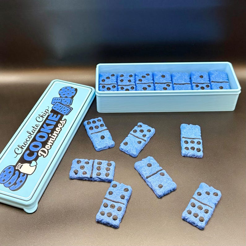 Puede incluir: Una lata y un recipiente azul claro contienen domin&oacute;s con forma de galleta. Los domin&oacute;s son azules con detalles de chispas de chocolate marr&oacute;n oscuro. La lata dice "Chocolate Chip Cookie Dominoes" en texto blanco y azul.