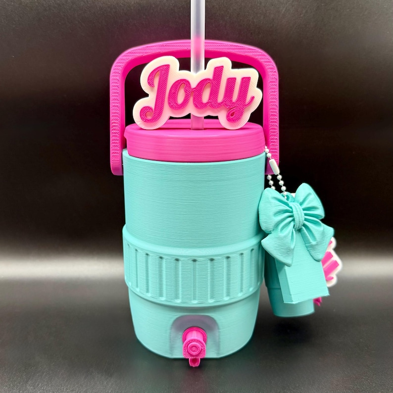 May include: A teal and pink water cooler with a handle and the name "Jody" on top. It has a pink lid, a teal body, and a pink spigot. A teal bow and tag hang from the side.