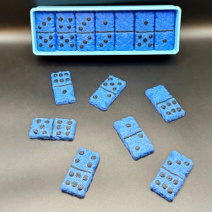 Puede incluir: Juego de domin&oacute; azul con puntos negros, presentado en un recipiente azul claro y disperso sobre una superficie oscura. Los domin&oacute;s tienen un aspecto texturizado. Un juego cl&aacute;sico para todas las edades.