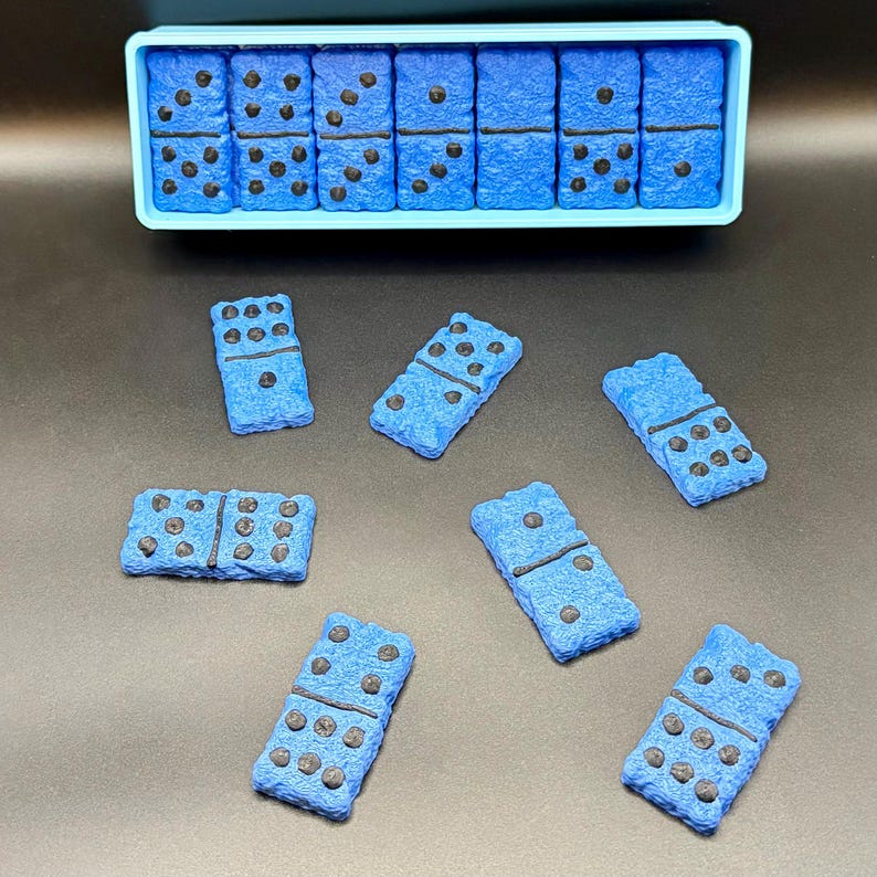Puede incluir: Juego de domin&oacute; azul con puntos negros, presentado en un recipiente azul claro y disperso sobre una superficie oscura. Los domin&oacute;s tienen un aspecto texturizado. Un juego cl&aacute;sico para todas las edades.
