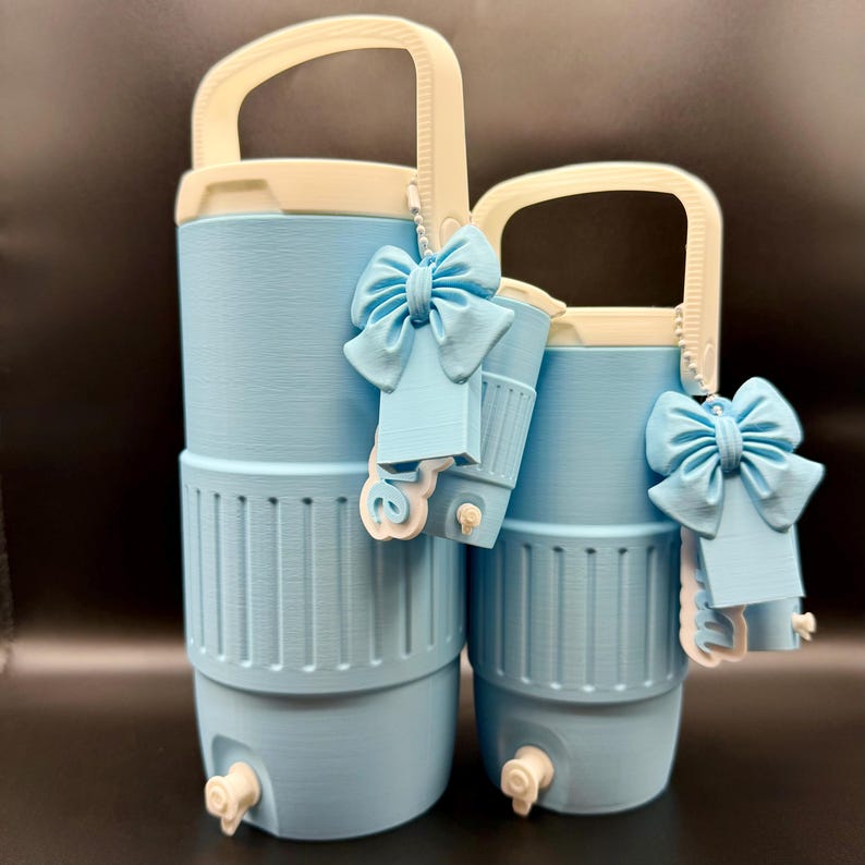 May include: Two light blue water coolers with white handles and accents. Each cooler has a decorative blue bow and a small tap. The coolers are cylindrical and appear to be made of plastic.