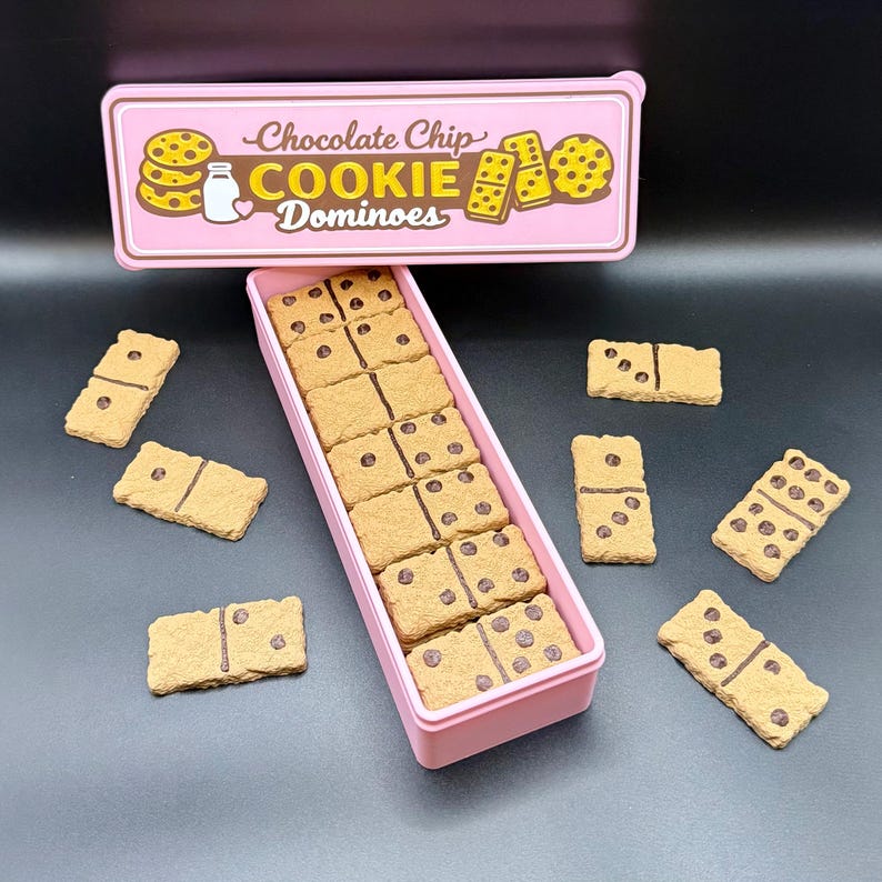 Puede incluir: Una caja y tapa rosas con la inscripci&oacute;n "Chocolate Chip Cookie Dominoes". La caja contiene domin&oacute;s con forma de galleta con detalles de chispas de chocolate. Varios domin&oacute;s est&aacute;n esparcidos sobre una superficie oscura.