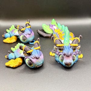 May include: Four colourful dragon figurines with iridescent blue, purple, and green scales. Each dragon has yellow accents, black horns, and green eyes. The dragons are lying down on a dark surface.