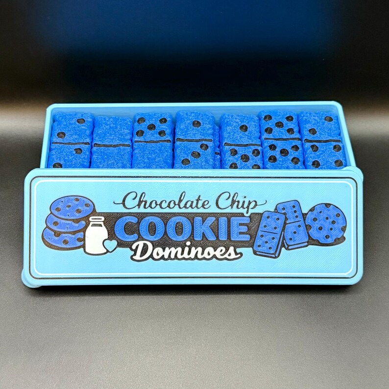 Puede incluir: Una caja azul claro de domin&oacute; con tem&aacute;tica de galletas con chispas de chocolate. La caja tiene las palabras "Chocolate Chip COOKIE Dominoes" e ilustraciones de galletas, domin&oacute; y una botella de leche. Los domin&oacute;s son azules con puntos negros.