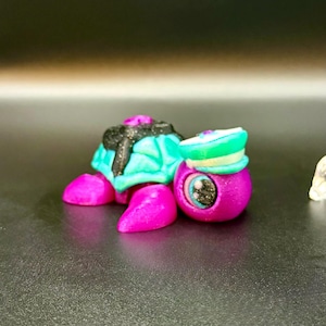May include: A whimsical, colorful turtle figurine with a teal shell, pink feet, and a black and glittery top. Next to it is a purple figurine with a green and white hat and a detailed eye. The figurines are on a dark surface.