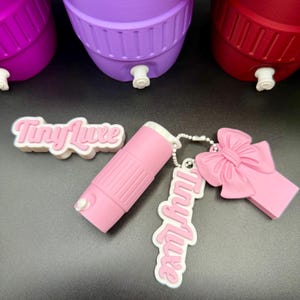 May include: A collection of pink accessories, including a lipstick-shaped item, a bow-shaped charm, and a keychain with the text "Tiny Luxe" in a cursive font. The items are set against a backdrop of colorful containers.