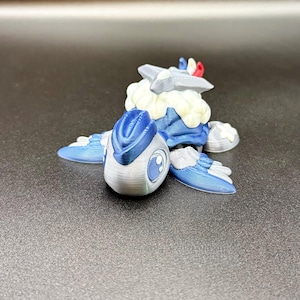 May include: A 3D-printed figurine of a stylized creature, in blue and white. The figure has a rounded head with a large eye, wings, and a cloud-like structure on its back. The figurine is resting on a dark surface.