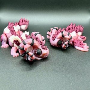 May include: Three handcrafted fantasy dragon figurines in shades of pink, white, and burgundy. The dragons have large, dark eyes and are detailed with black accents. The figurines are lying on a dark grey surface.