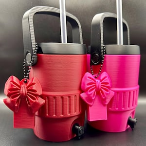 May include: Two insulated tumblers, one red and one pink, with black handles and lids. Each tumbler has a white straw and a matching bow-shaped charm. The tumblers have a textured surface and a small spout near the base.