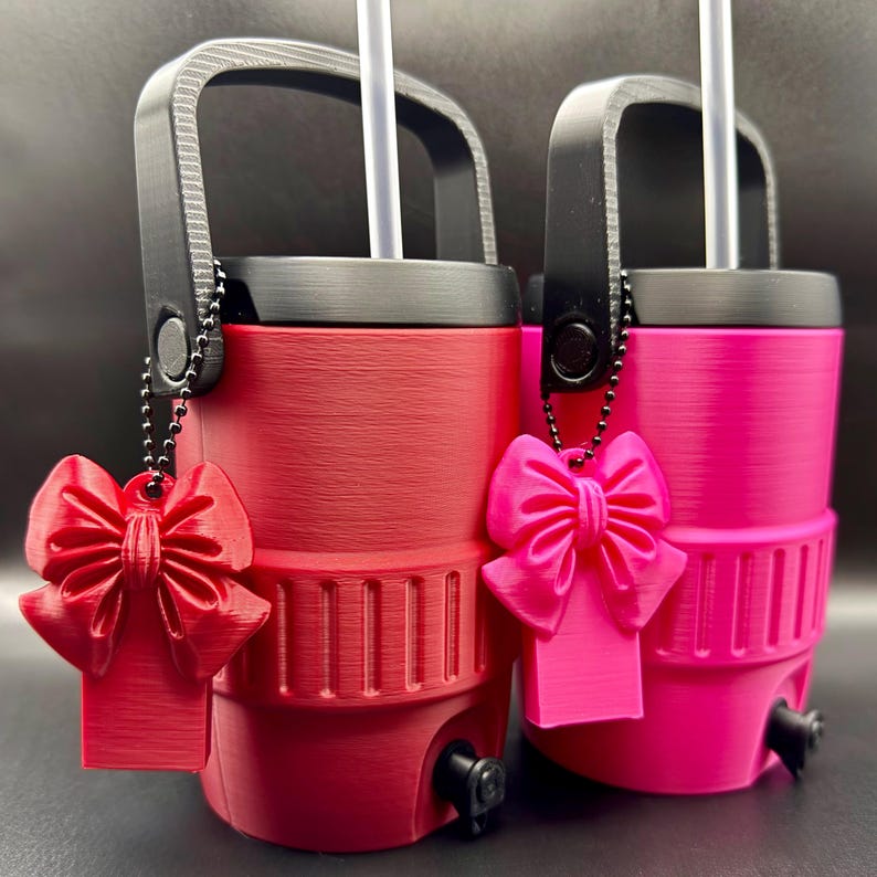 May include: Two insulated tumblers, one red and one pink, with black handles and lids. Each tumbler has a white straw and a matching bow-shaped charm. The tumblers have a textured surface and a small spout near the base.