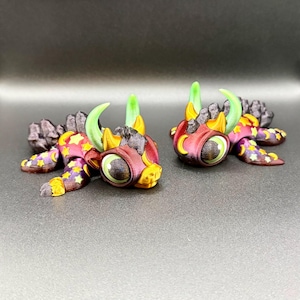 May include: Two colourful, whimsical figurines. Each has a maroon body, large green-eyed face, and yellow and green horns. The bodies are decorated with yellow stars and purple crescent moons. The backs are dark grey and textured.