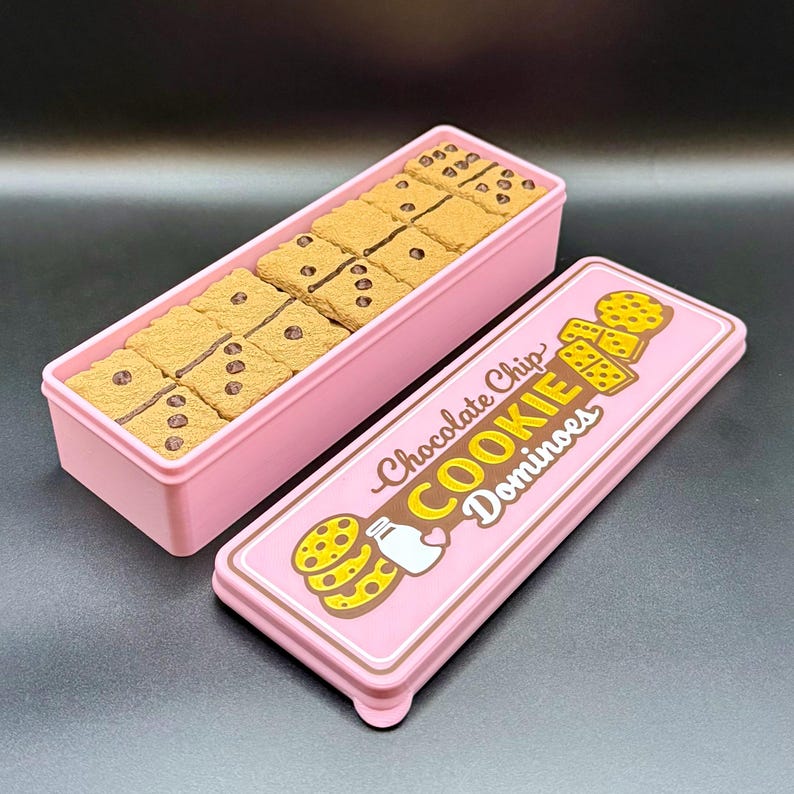 Puede incluir: Una caja rectangular rosa con tapa, que contiene domin&oacute;s con forma de galleta. La tapa presenta el texto "Chocolate Chip COOKIE Dominoes" con ilustraciones de galletas y leche. Los domin&oacute;s son de color marr&oacute;n claro con chispas de chocolate.