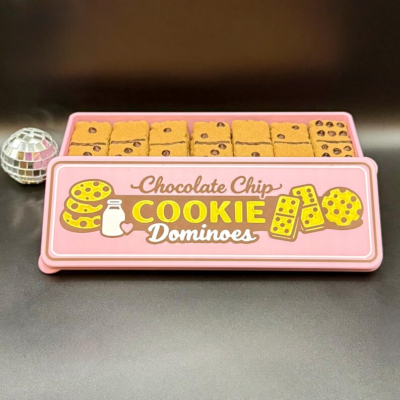 Puede incluir: Una caja rosa de domin&oacute; de galletas con chispas de chocolate. Los domin&oacute;s son marrones con detalles de chispas de chocolate. La caja tiene las palabras "Chocolate Chip COOKIE Dominoes" en una fuente l&uacute;dica, junto con ilustraciones de galletas y leche.