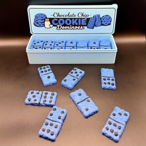 Puede incluir: Una caja azul claro de "Chocolate Chip Cookie Dominoes" con una tapa a juego. La caja contiene domin&oacute;s azules con detalles de chispas de chocolate. Varios domin&oacute;s est&aacute;n esparcidos alrededor de la caja sobre una superficie marr&oacute;n.
