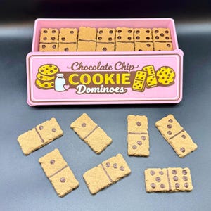 Puede incluir: Una caja rosa de domin&oacute;s de galletas con chispas de chocolate con el texto "Chocolate Chip Cookie Dominoes". Los domin&oacute;s son de color canela con chispas de chocolate marr&oacute;n. Algunos domin&oacute;s est&aacute;n fuera de la caja.