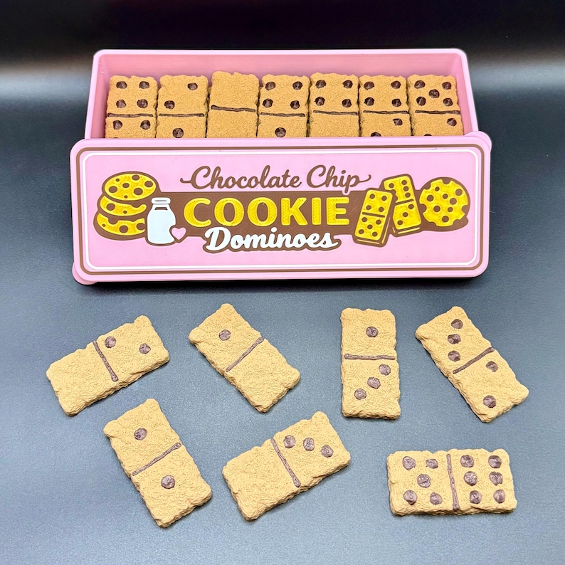 Puede incluir: Una caja rosa de domin&oacute;s de galletas con chispas de chocolate con el texto "Chocolate Chip Cookie Dominoes". Los domin&oacute;s son de color canela con chispas de chocolate marr&oacute;n. Algunos domin&oacute;s est&aacute;n fuera de la caja.