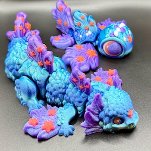 May include: A collection of colourful, 3D-printed fantasy creatures in shades of blue, purple, and orange. The creatures have a scaled texture and star-shaped accents, with a detailed eye and fin-like structures. The pieces are arranged on a dark surface.