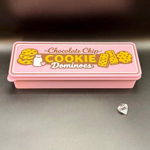 Puede incluir: Una caja rectangular rosa con tapa que presenta el texto "Chocolate Chip COOKIE Dominoes" en marr&oacute;n y amarillo. La tapa tiene ilustraciones de galletas y una botella de leche. Una gema transparente facetada se encuentra a un lado.