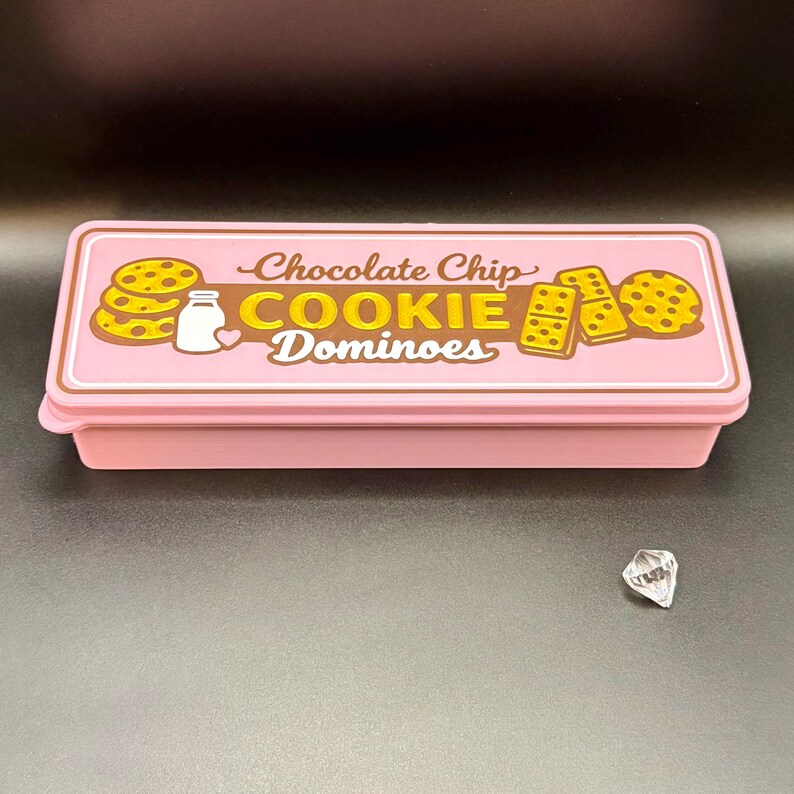 Puede incluir: Una caja rectangular rosa con tapa que presenta el texto "Chocolate Chip COOKIE Dominoes" en marr&oacute;n y amarillo. La tapa tiene ilustraciones de galletas y una botella de leche. Una gema transparente facetada se encuentra a un lado.