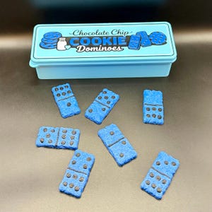 Puede incluir: Una caja azul claro con las palabras "Chocolate Chip Cookie Dominoes" e ilustraciones de galletas y domin&oacute;s. Varios domin&oacute;s azules con puntos negros est&aacute;n esparcidos alrededor de la caja.
