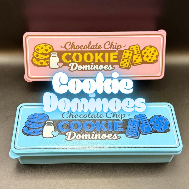 Puede incluir: Dos cajas rectangulares con las palabras "Cookie Dominoes". La caja superior es rosa con ilustraciones de galletas y domin&oacute;s, y el texto "Chocolate Chip". La caja inferior es azul con ilustraciones y texto similares.