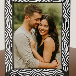 May include: A rectangular photo frame with a black and white zebra print border. The frame holds a photograph of a couple embracing. The frame is supported by two small, black stands.