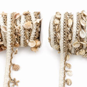 May include: Two spools of decorative trim featuring a white woven border and a tan mesh center. The left spool is adorned with small seashells and starfish, while the right spool is densely decorated with numerous small, smooth, light-colored seashells.