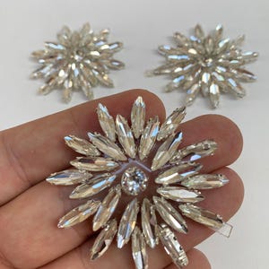 Può includere: Tre applicazioni decorative circolari con cristalli trasparenti a taglio marquise disposti a raggiera. Ogni pezzo presenta un cristallo rotondo centrale circondato da più strati di pietre appuntite e sfaccettate che riflettono la luce.