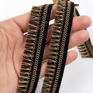 May include: A black and gold fringe trim featuring a central woven band with a cross-stitch pattern, flanked by metallic gold-toned fringe edges. The decorative ribbon displays a high-contrast color scheme with a textured finish throughout.