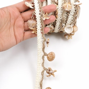 May include: A spool of decorative trim featuring a white woven fabric band paired with a natural tan jute cord. Attached to the trim are small, dangling charms shaped like various seashells and starfish in shades of beige, brown, and white.