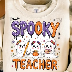 Puede incluir: Sudadera color crema con las palabras "SPOOKY TEACHER" en morado y naranja, con un diseño de murciélago y telaraña. Tres fantasmas de dibujos animados con coronas de flores aparecen debajo del texto, sosteniendo un lápiz, una calabaza y libros.