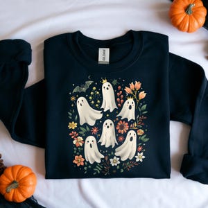 May include: A navy blue sweatshirt featuring a whimsical Halloween design. The graphic includes several white ghost figures with crowns, surrounded by colorful flowers, leaves, and a bat. The sweatshirt is a cozy, long-sleeved garment.