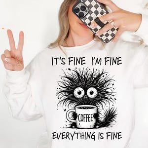 Puede incluir: Sudadera blanca con el texto "IT'S FINE I'M FINE EVERYTHING IS FINE" y una ilustración de dibujos animados de una criatura negra y peluda sosteniendo una taza de café. La taza tiene la palabra "COFFEE". También se ve una gorra beige y un teléfono con una funda a cuadros en blanco y negro.