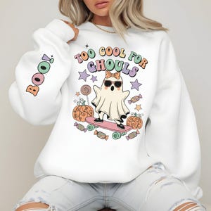 Puede incluir: Sudadera blanca con un diseño temático de Halloween. El diseño presenta un fantasma con gafas de sol y un lazo, montando en monopatín, con el texto "TOO COOL FOR GHOULS". La manga tiene la palabra "BOO" en colores pastel.