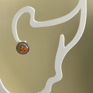 Può includere: Un singolo orecchino a lobo con castone color argento. L'orecchino presenta un design colorato e maculato con sfumature arancioni, gialle, verdi e rosa. L'orecchino è esposto su un espositore bianco astratto.